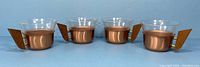 Set of four Schott Mainz Jenaer glass cups with copper cradles and angled teak wood handles shown from a front view.