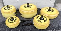 Full set of five yellow cookware pieces with black sunburst lids and black handles, arranged outdoors on a pavement against a white wall.
