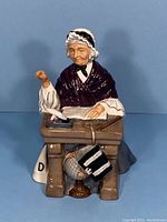 Front view of the Royal Doulton Schoolmarm figurine showing the elderly woman seated at a school desk with an inkwell, open book, and quill.