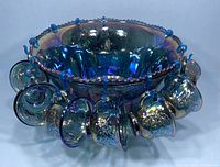Front angled photo of blue carnival glass punch bowl with attached hooks and cups hanging on them, showing detailed floral pattern and iridescent finish.