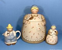 Three ceramic figurines including a cream-colored cookie jar woman holding a spoon, creamer jug man with straw hat and pipe, and sugar bowl woman holding kitten.