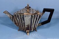 Front left side view showing geometric ridged silver plated body, black angular handle, spout, and decorative finial on lid.