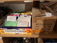 Photo showing assortment of CD/DVD sleeves and cases, boxes labeled Memorex, 100-count packs including colorful plastic slim cases and paper sleeves.