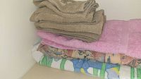 Stack of assorted bath towels and blankets including beige towels, a pink towel, and patterned blankets with floral and nature themes.