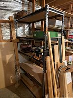 Metal storage shelf unit with four shelves, against a wall with various items around it, empty shelves.