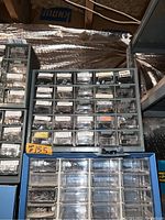Front view of three plastic organizer drawer units with assorted small hardware items like screws, nails, hooks, and brackets sorted in clear drawer compartments with labels.