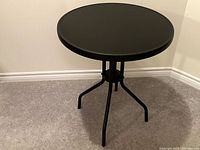 Front and top view of a black round patio table with four angled legs on carpeted floor near wall corner.