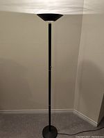 Full view of the floor lamp standing against a wall showing its height, black metal pole, circular base, and bowl-shaped shade.