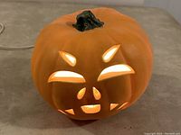 Orange mock pumpkin with carved Jack O Lantern face lit from inside, connected to electrical cord, showing front expression and stem detail.