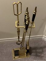 Front view of brass fireplace tool set with tools neatly arranged on stand showing black-handled fire poker, brush broom, shovel, and log tongs