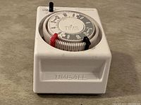 White plastic Intermatic Time-All mechanical timer with round 24-hour dial, black and red trippers, embossed TIMEALL label, side view showing dial face.