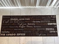 Rectangular coffee-themed kitchen floor mat with text related to coffee types and drinks, placed on a tile floor.