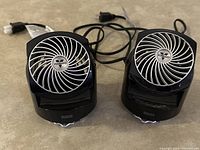 Front view of two black Vornado Flippi V6 fans placed side by side, showing circular front grille with Vornado logo and control knobs labeled off, low, high speeds.