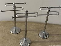 Three chrome metal towel stands with S-shaped arms standing upright on a flat surface, showing overall condition and design.