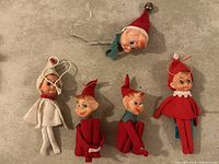 Photo showing five elf Christmas tree ornaments with plastic heads and fabric bodies, including one with a white outfit, three with red outfits and varying collars, and one elf head with a red hat with a silver bell.