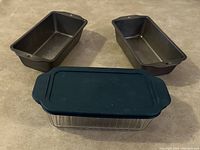 Two steel loaf pans with rectangular shape and dark finish, placed side by side, and one clear rectangular glass container with a dark blue rubber lid on top.