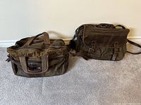 Two Old West style olive green leather bags on carpet floor, one larger luggage bag and one smaller carry-on crossbody bag.