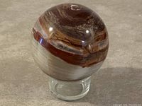 Close up of the brown quartz sphere mineral stone on a clear glass pedestal, showing natural horizontal banding and polished surface.