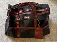 Full front view of the vintage Samsonite duffle gym bag showing black fabric and red leather handles, zipper, and trim, with an ID tag attached.