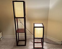 Front view of two Asian style floor lamps, dark wooden frames with beige fabric shades, lit to show light diffusion. Taller lamp has shelf below shade, smaller lamp is shorter with storage space at base.