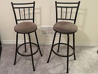 Front view showing two identical black steel four-legged swivel barstools with cushioned gray fabric seats and high backs, placed on carpeted floor against white wall.