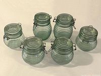 Photo showing the full set of six green tinted glass food storage containers with metal latches. Four shorter and two taller containers are displayed with a ribbed texture and airtight metal clamps.