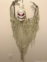 Skeleton Reaper Hanging Decoration with glowing red eyes, raised arms holding chains, and tattered fabric draping. Mounted on wall showing full figure.