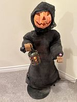 Full view of the talking Halloween Jack O Lantern scarecrow in black hooded robe holding a light-up lantern. Pumpkin face is unlit.