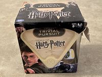 Front view of the boxed Trivial Pursuit World of Harry Potter Edition game showing the triangular game cartridge inside with Harry Potter-themed graphics. Box has a small tear on the top right corner.