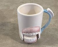 White ceramic rinse cup with toothpaste-shaped handle and movable pink-and-white dentures attached to the front.