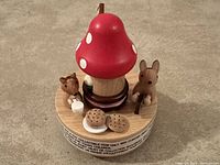Front view of the wooden music box showing the red mushroom house with white spots, rabbit and squirrel figures, and small wooden cookies and mug details on the wooden base.