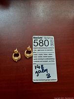 Pair of 14K gold earrings on a red surface next to a MaxSold label noting 14K gold material and signs of wear