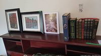 Books and framed pictures on wooden shelf, including classic books and three framed photographs in black and white frames.