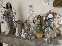 Full view of ten angel figurines arranged on a shelf including stone, wood, glass, and fabric types in various sizes and styles
