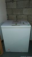 Front view of white Frigidaire chest freezer against a concrete wall showing the freezer's closed lid and a control panel with documents taped to the top.