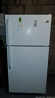 Front view of white Frigidaire top freezer refrigerator showing both freezer and fridge doors with handles and label stickers attached.