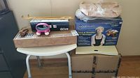 Overall front view of the lot showing various boxed and unboxed items on and around a white bathroom stool including Roho Mosaic seat cushion, Mobb pressure mattress, Quantum neck massager, Drive grab bar, Homedics massager, two wooden back scratchers, and glasses holder.