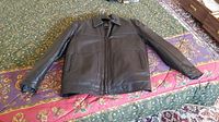 Black Pelle Cuir leather jacket laid flat on a patterned bedspread, showing front view with sleeves extended.