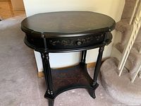 Front view of black oval side table showing painted floral decorations, drawer, and lower shelf.