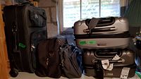 View of two silver hard shell expandable wheeled suitcases stacked with black medium wheeled cloth suitcase and small dark backpack in front. Small travel accessories bags also visible in front of suitcases.