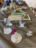 Photo showing six Swarovski figurines on a table including a crystal horse on a mirrored base, rabbit with pink ears, koala bear, turtle dove, hummingbird, and butterfly on leaf.