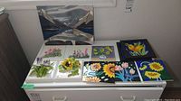 Wide view showing nine ceramic tiles arranged on a white surface, each with distinct floral or landscape designs, colorfully painted.