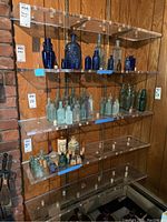 Five clear acrylic shelves mounted on wall, each holding various vintage glass bottles in blue, green, and clear glass.