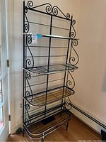 Full view of the black metal four tier bakers rack with gold trim standing near a door.