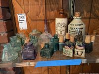 Front view of the bottles, crocks, cork tops on glass shelf with wooden background showing variety of shapes, colors, and sizes