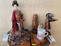 Full view of lot including Chinese doll, carved stone vase, wooden tiki figure, fiddler man figurine, Hercules figure, and mug.