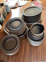 Photo shows multiple stacks of stoneware dinnerware including oval plates, round plates, bowls, and mugs with a dark speckled finish.