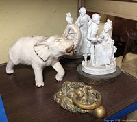 Elephant figurine with raised curled trunk and coin slot on the back, white porcelain couple figurine dressed in vintage attire on round base with gold trim, and brass lion door knocker showing signs of wear.