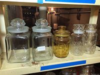 Photo showing all five glass canisters in a row on shelf, including two large clear square canisters, amber canister, and two smaller patterned canisters with lids.