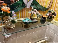 Glass shelf displaying three miniature floral arrangements and one cloisonne pot with lid and handle in front of stained glass wall.